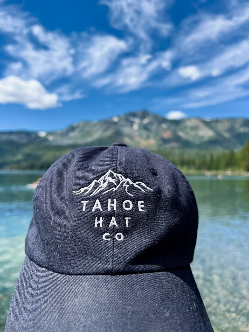 Close view of white mountain embroidery on a navy cap against a lake and mountain backdrop